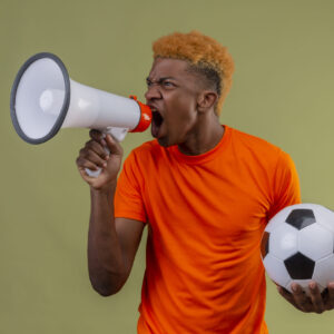 young african american boy wearing orange t-shirt holding soccer ball shouting to megaphone with angry expression on face standing over green background
