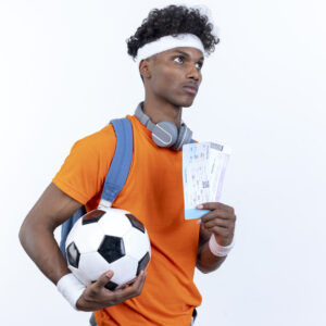 looking at side young afro-american sporty man wearing headband and wristband holding ball and tickets isolated on white background